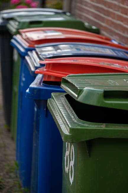An outdoor scene depicting a large pile of cardboard boxes, primarily containing fresh fruit packaging, stacked on a paved surface. The boxes are colourful, with labels showing images and text related